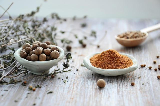 Close-up of whole black peppercorns from Zanzibar in a small wooden scoop with a brass pepper grinder and scattered peppercorns on a dark stone surface